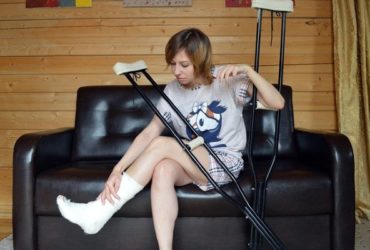 A blond woman sitting on a couch with crutches and her foot in a cast