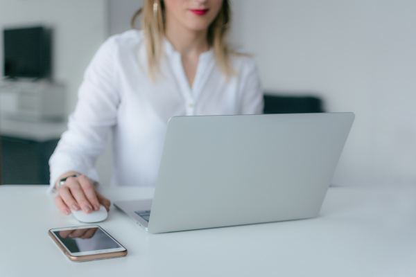 A blonde woman wearing a white top sitting at white desk using a laptop mouse and looking at laptop
