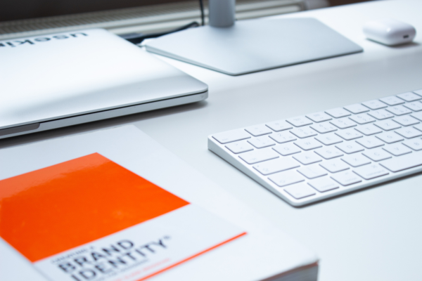 Table with desktop computer, wireless keyboard and orange notebook with brand identity written on it.